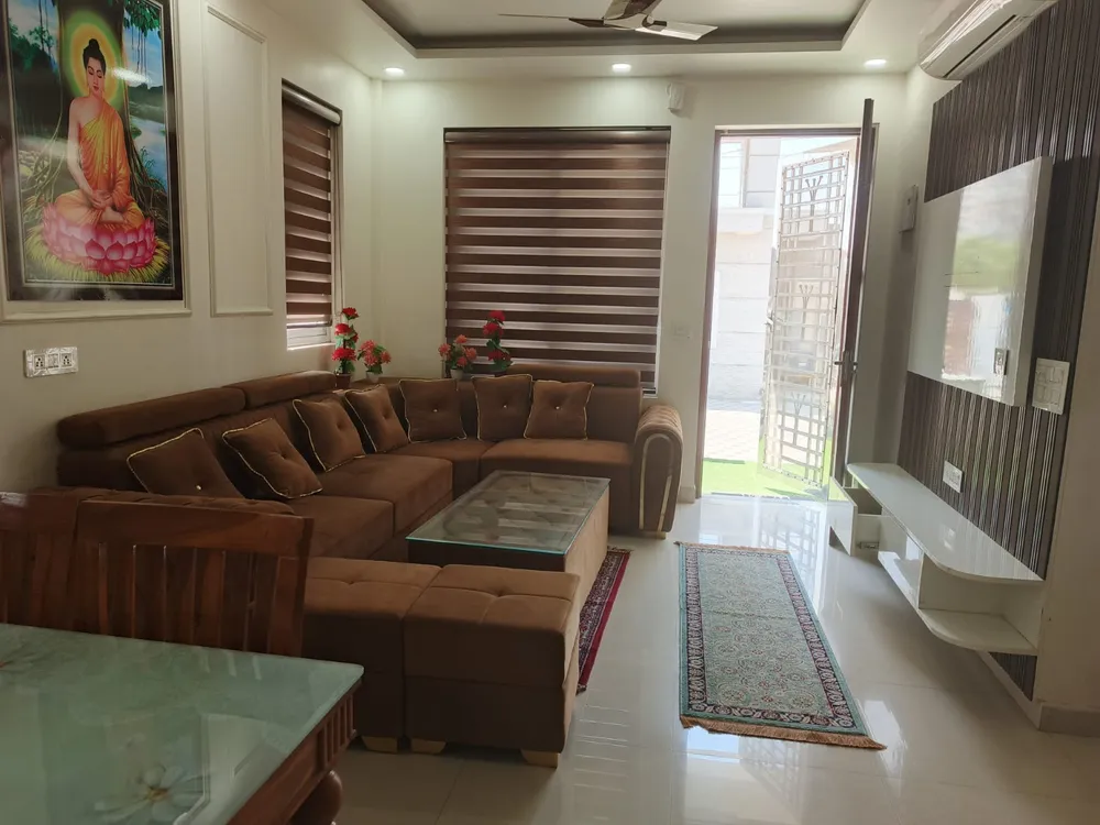 Living room of a 5 BHK villa in Madhuvan Residency with an L-shaped sofa, glass-top table and panelled TV wall, Bhiwadi