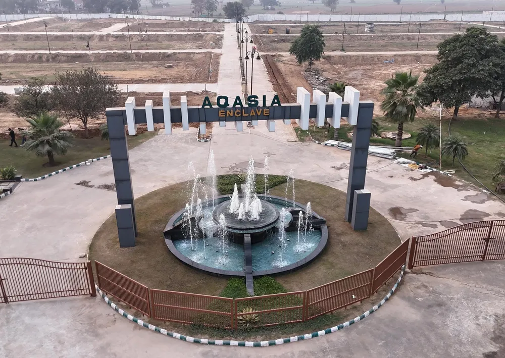 The Aqasia Enclave entrance arch and fountain seen from above, with the kerbed avenue and graded plots behind, Alwar Bypass Road, Bhiwadi
