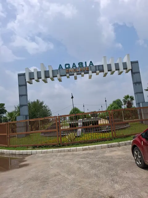The Aqasia Enclave entrance arch on the Bhiwadi mega-highway, project name in green lettering above the gate, with a planted roundabout behind the boundary railing