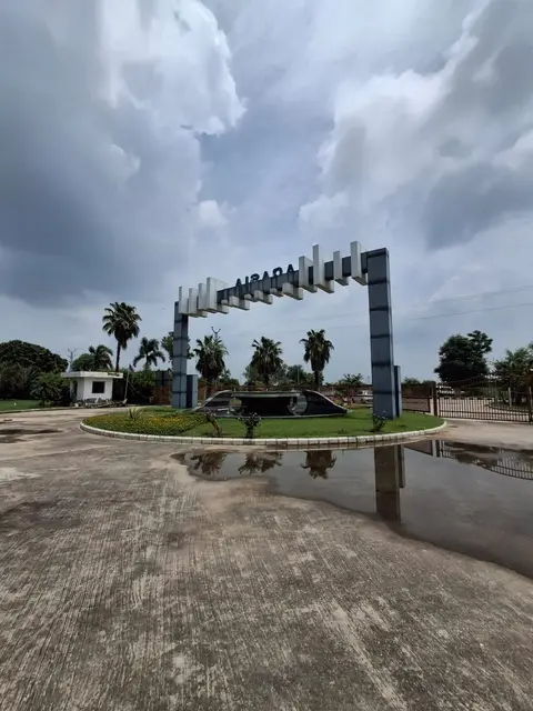 The Aqasia Enclave gate seen from inside the township under a monsoon sky, showing the water feature, guard cabin and boundary gate