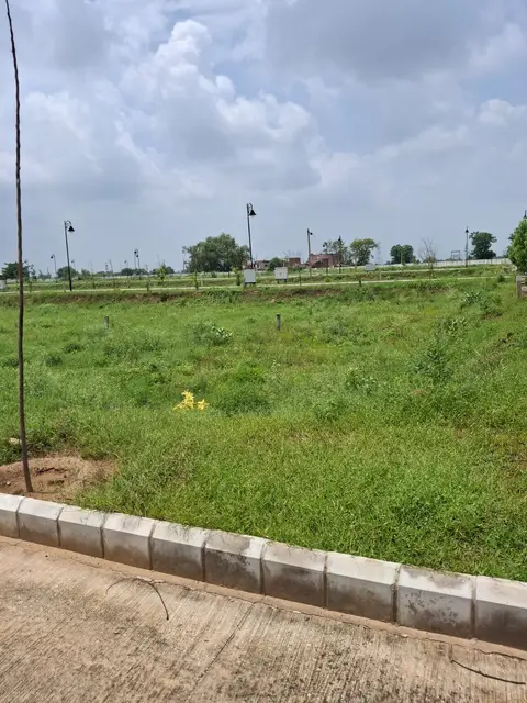 Vacant grassed plots at Aqasia Enclave with corner markers and a kerbed road, street lights installed, village houses on the horizon