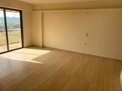 Empty living room with wood-finish floor tiles and a sliding door to the balcony in an Ashiana Tarang flat, Bhiwadi