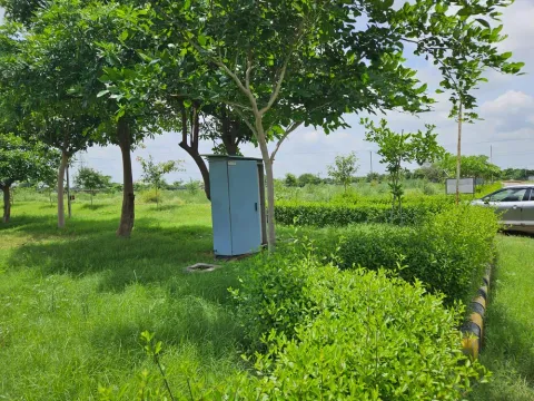 Planted green belt between plot rows at Krish Season, with an electricity distribution cabinet under the trees